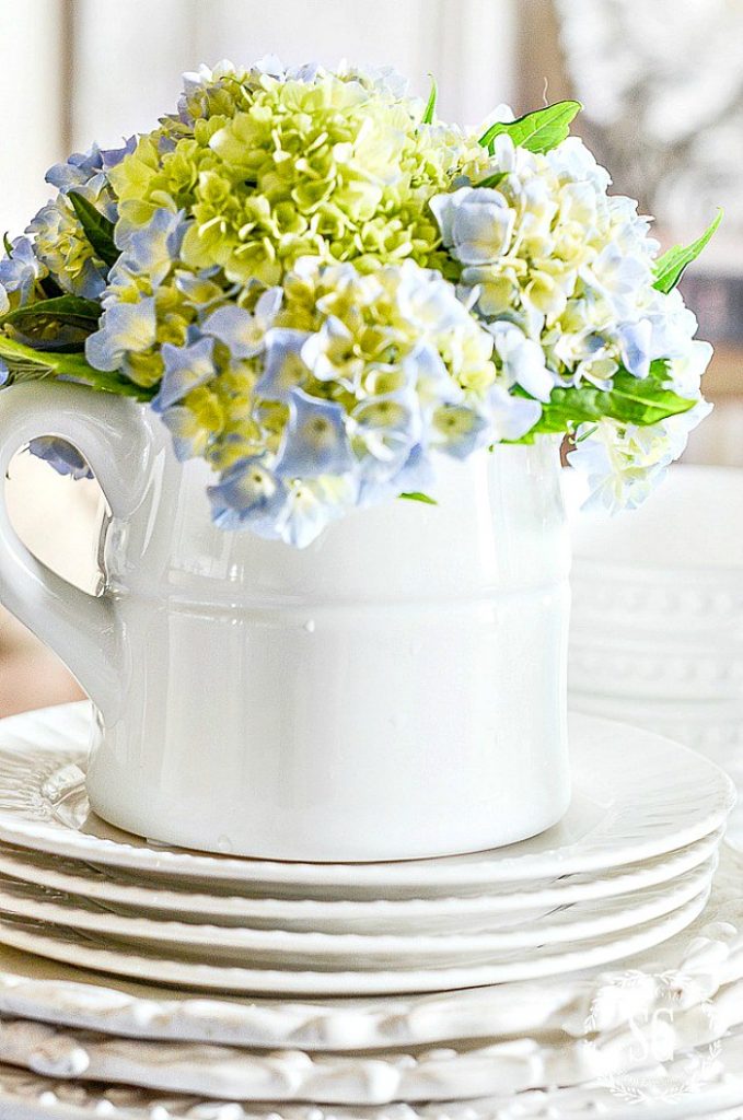 STOUT WHITE PITCHER WITH BLUE HYDRANGEAS IN IT ON A STACK OF WHITE PLATES