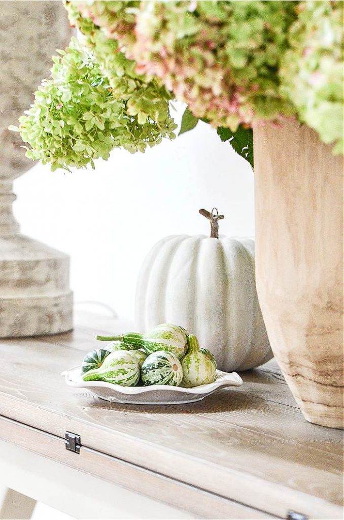 A FOYER TABLE DECORATED WITH HYDRANGEAS, A WHITE PUMPKIN AND A PLATE OF TINY GOURDS