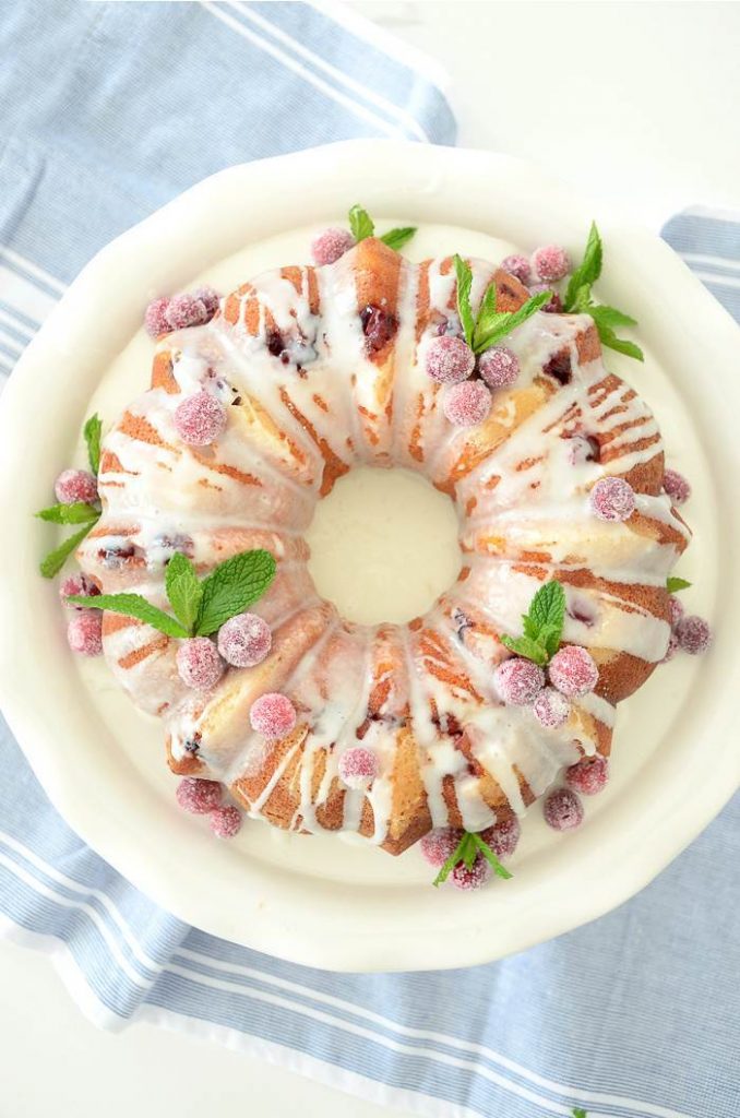 A CRANBERRY ORANGE BUNDT CAKE  DECORATED WITH SUGARED CRANBERRIES AND MINT LEAVES.