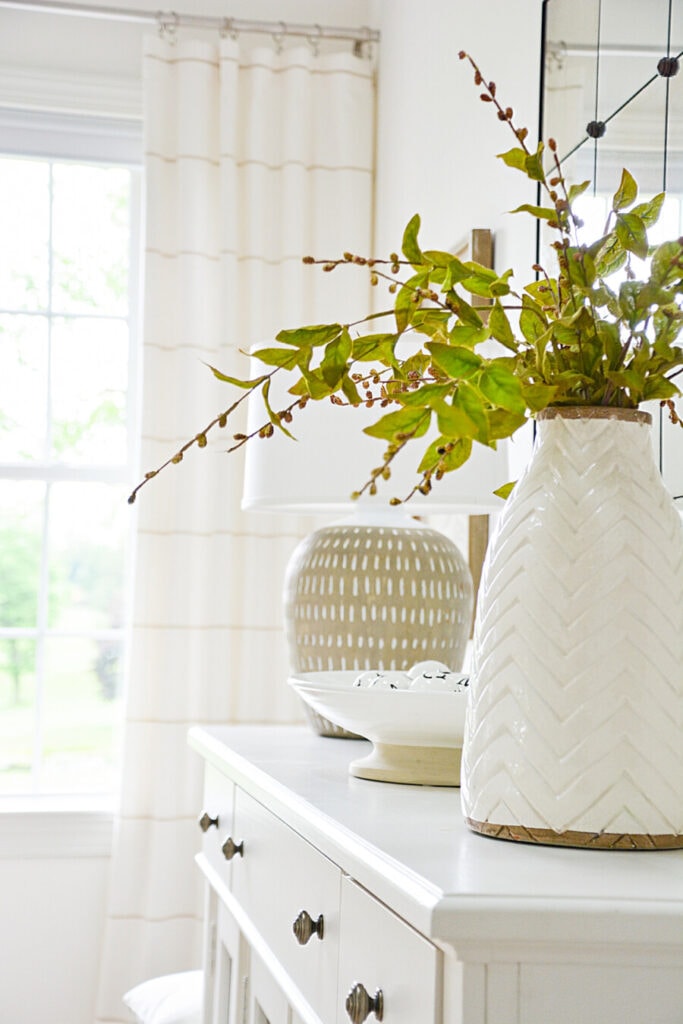 TIMELESS DECOR- WHITE BUFFET WITH A LAMP AND A VASE OF GREENS