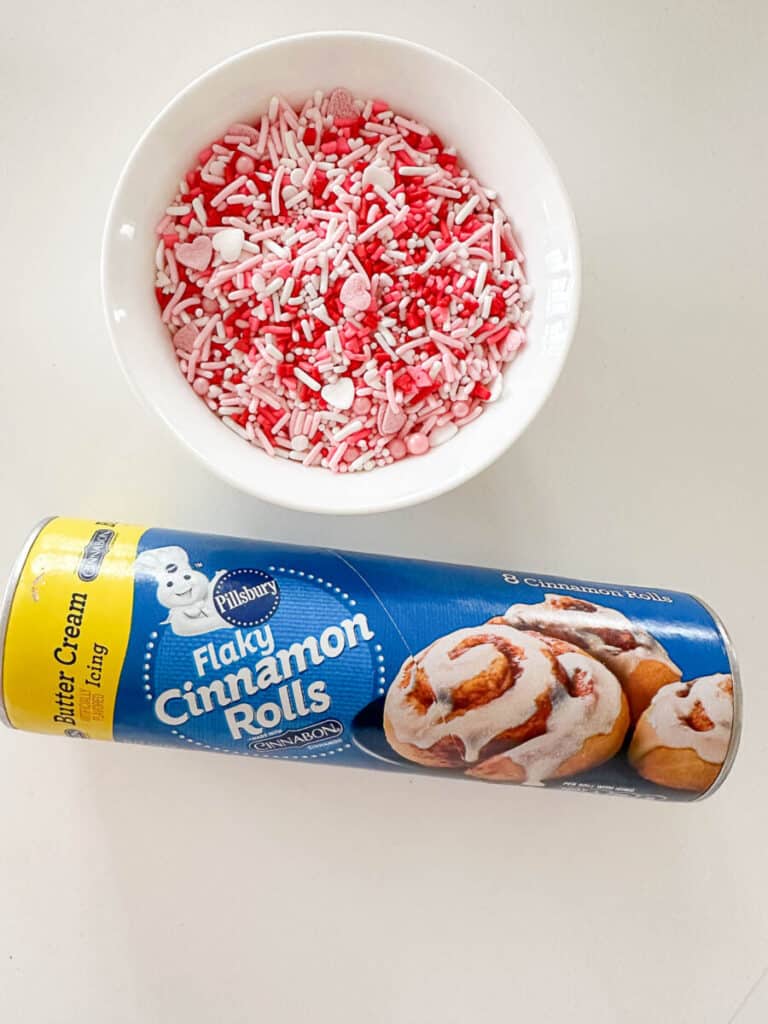 a small bowl of red, white and pink sprinkles and a can of Cinnamon Rolls
